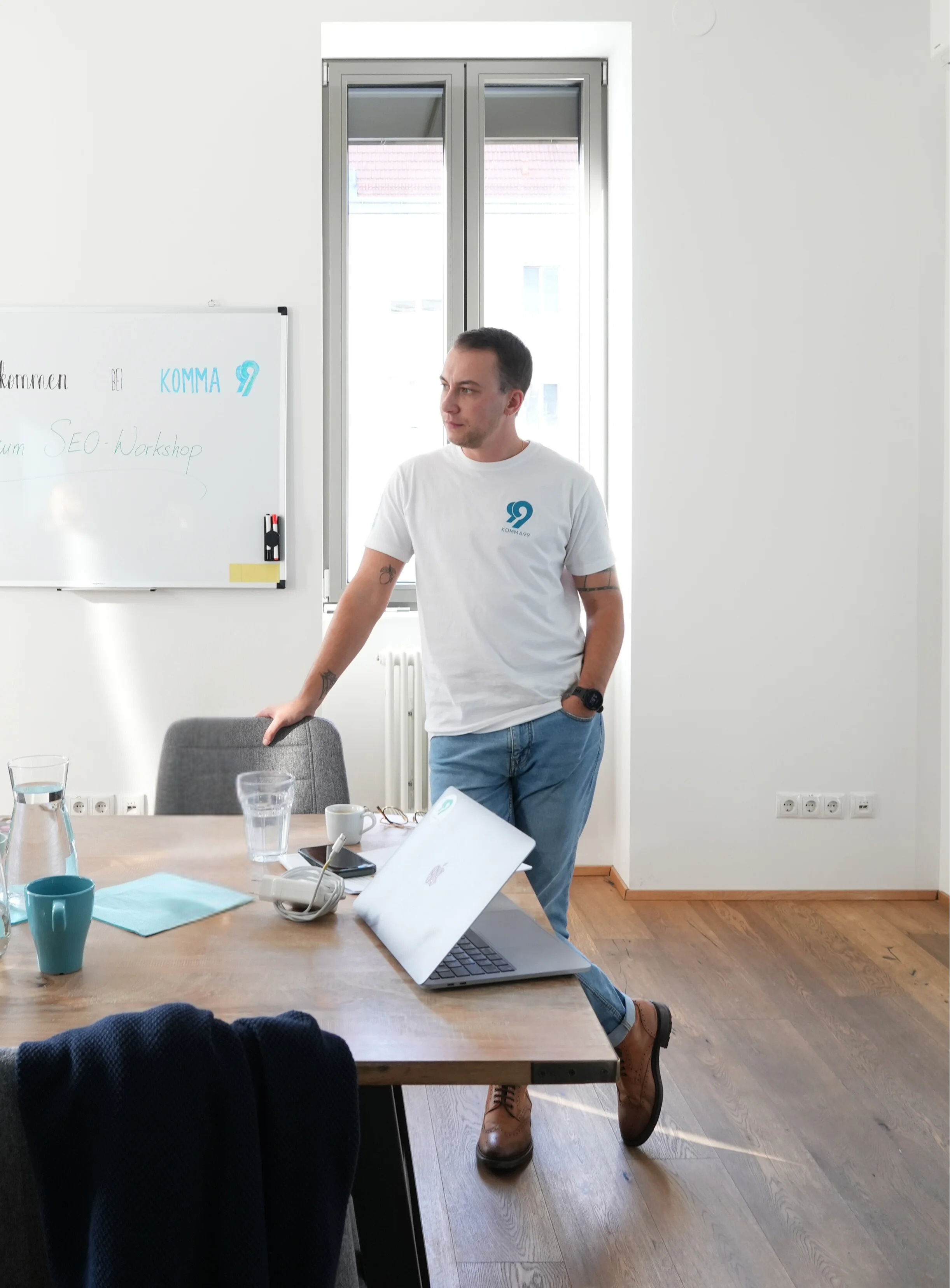 Ein Mann in einem weißen T-Shirt mit dem Logo von KOMMA99 steht entspannt an einem Konferenztisch in einem hellen Büro. Auf dem Tisch befinden sich ein Laptop, Gläser, eine Karaffe mit Wasser und Notizen. Im Hintergrund ist ein Whiteboard mit der Aufschrift „SEO-Workshop“ und ein großes Fenster mit Tageslicht zu sehen.