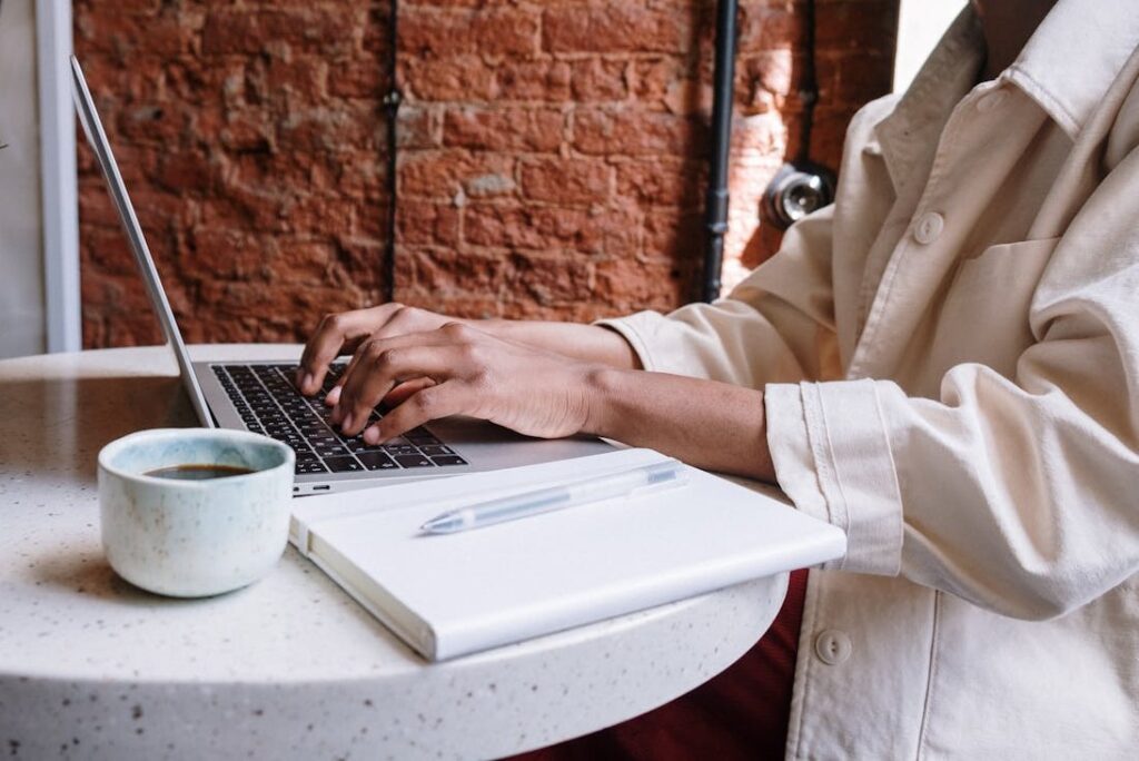 Content Marketing einfach erklärt 1 Person sitzt an einem Tisch und tippt auf einem Laptop, mit einem Notizblock und einer Tasse Kaffee daneben